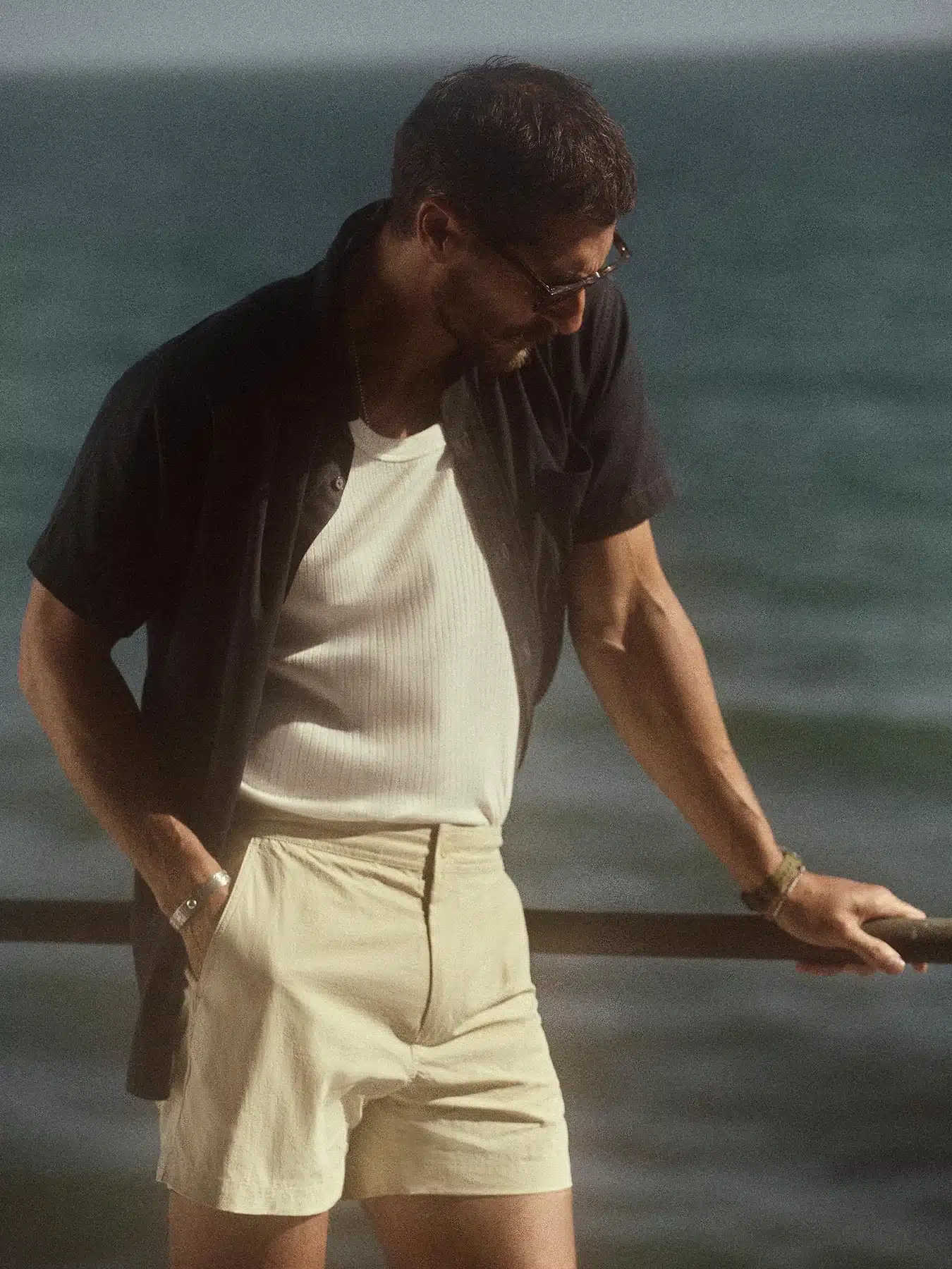 Man at the beach wearing an open camp collar shirt, white ribbed tee, and soft sand swim shorts