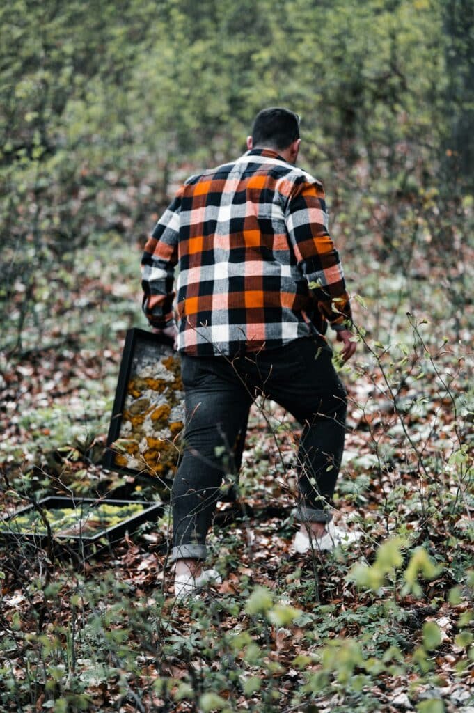 Man in an orange, white and black check flannel shirt walking through fallen leaves in a forest