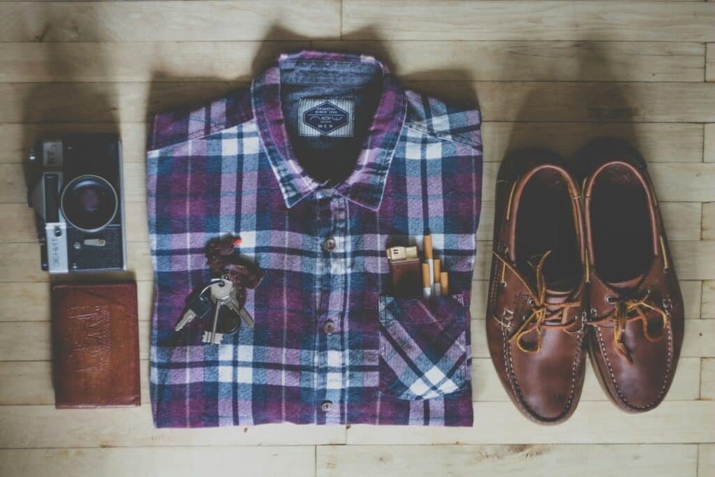 Flat lay of a plaid button-up shirt, brown boat shoes, a camera, keys and a passport