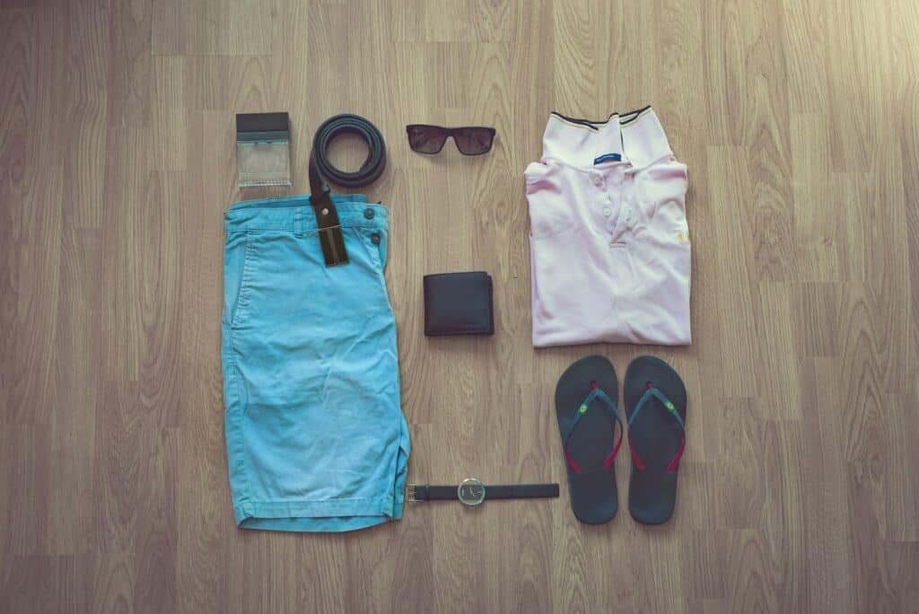 Flat lay of light blue shorts, a white polo, flip-flops, sunglasses, a belt and a wallet