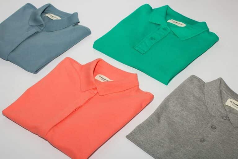 Four folded polo shirts in gray, green, coral and slate on a white surface