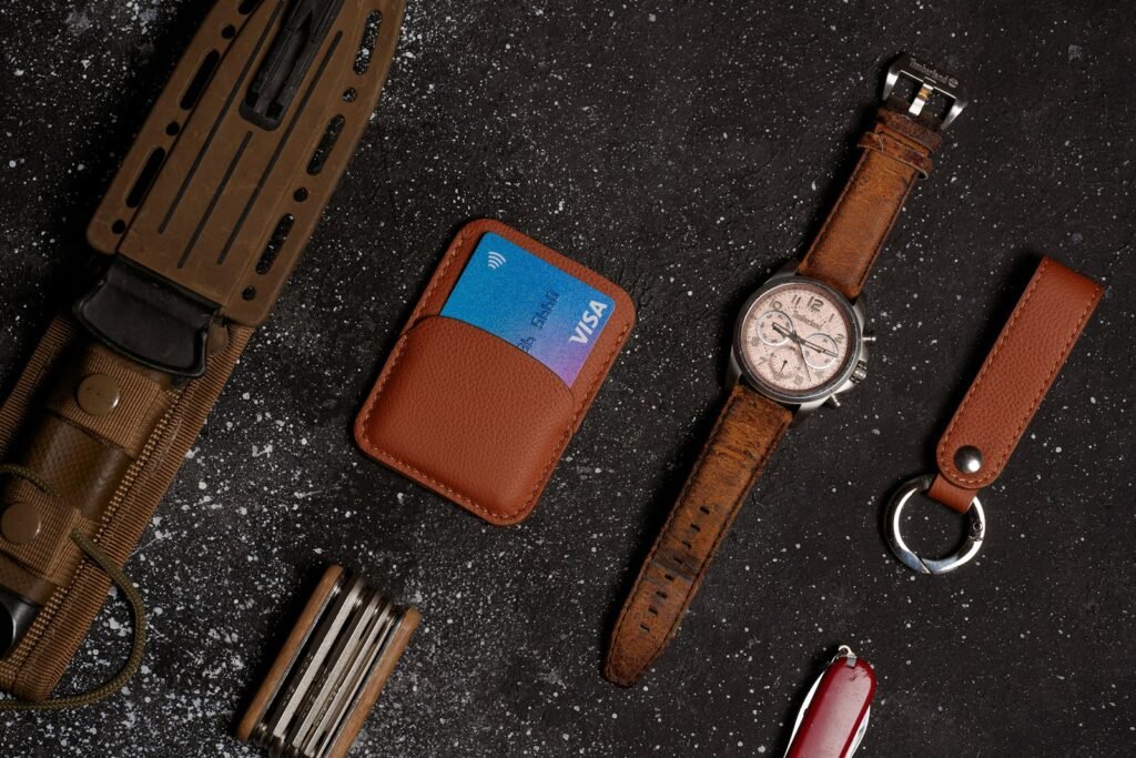 Leather card wallet, wristwatch and leather key fob laid out on a dark stone surface