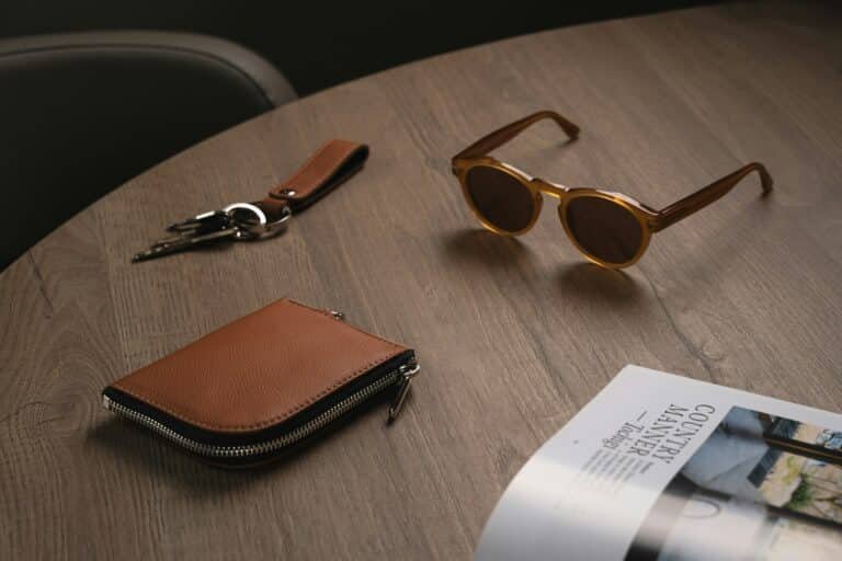 Brown leather zip wallet, keys and sunglasses on a round wooden table