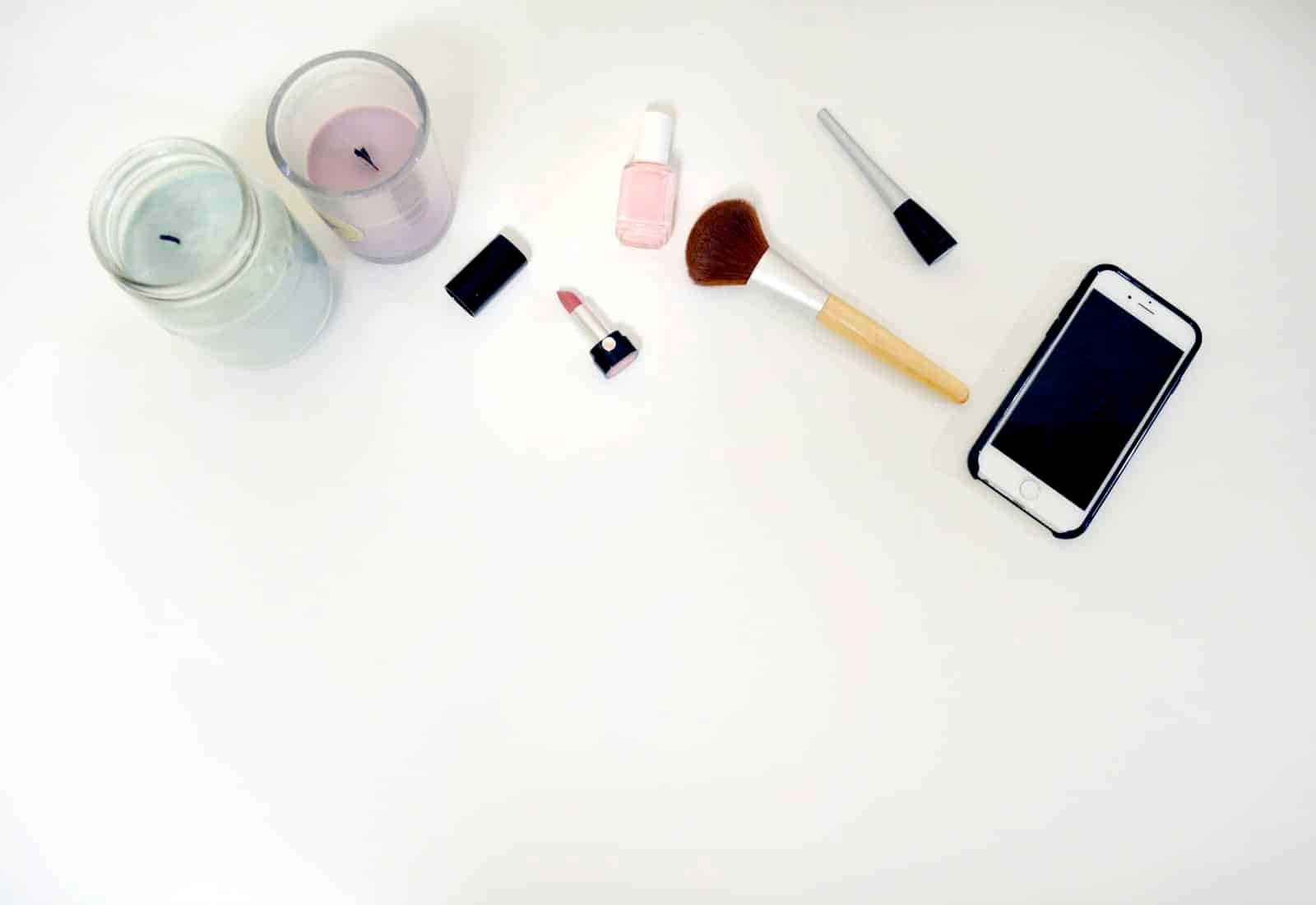 Makeup brushes, lipstick, nail polish, candles and a phone on a white surface