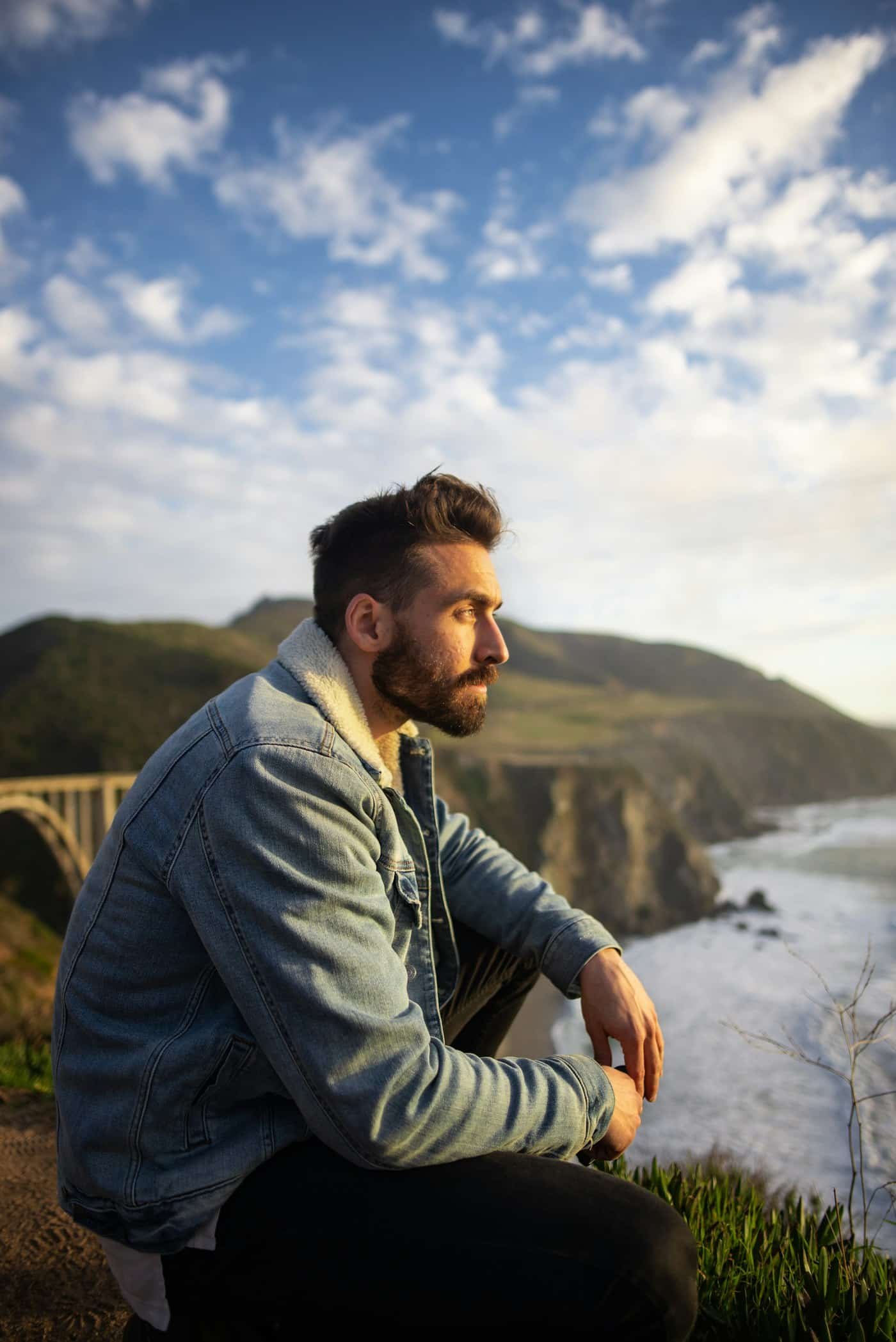 Man in a sherpa-lined denim jacket sitting on a grassy cliff above the ocean