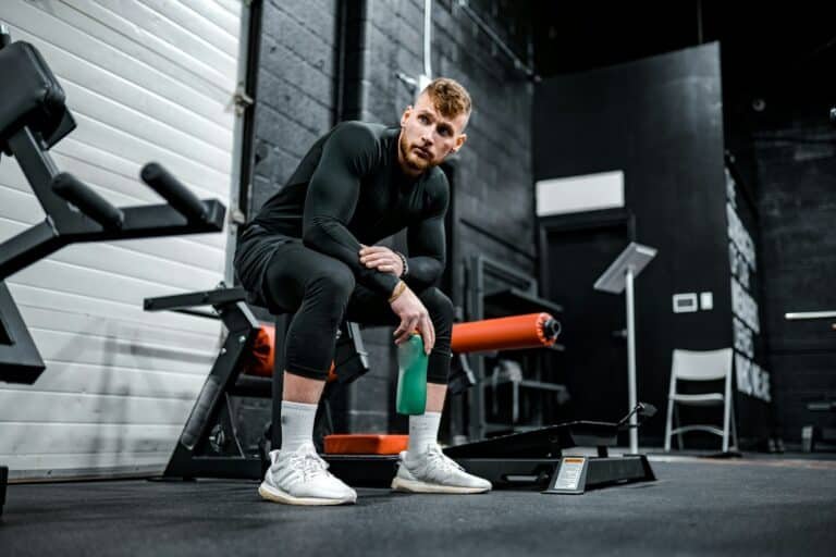 Man in a black long-sleeve top and tights resting on a gym bench with a water bottle