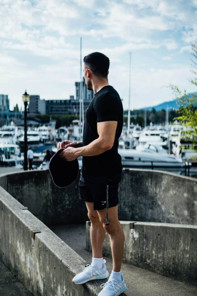 Man in a black tee and shorts holding a cap at a marina