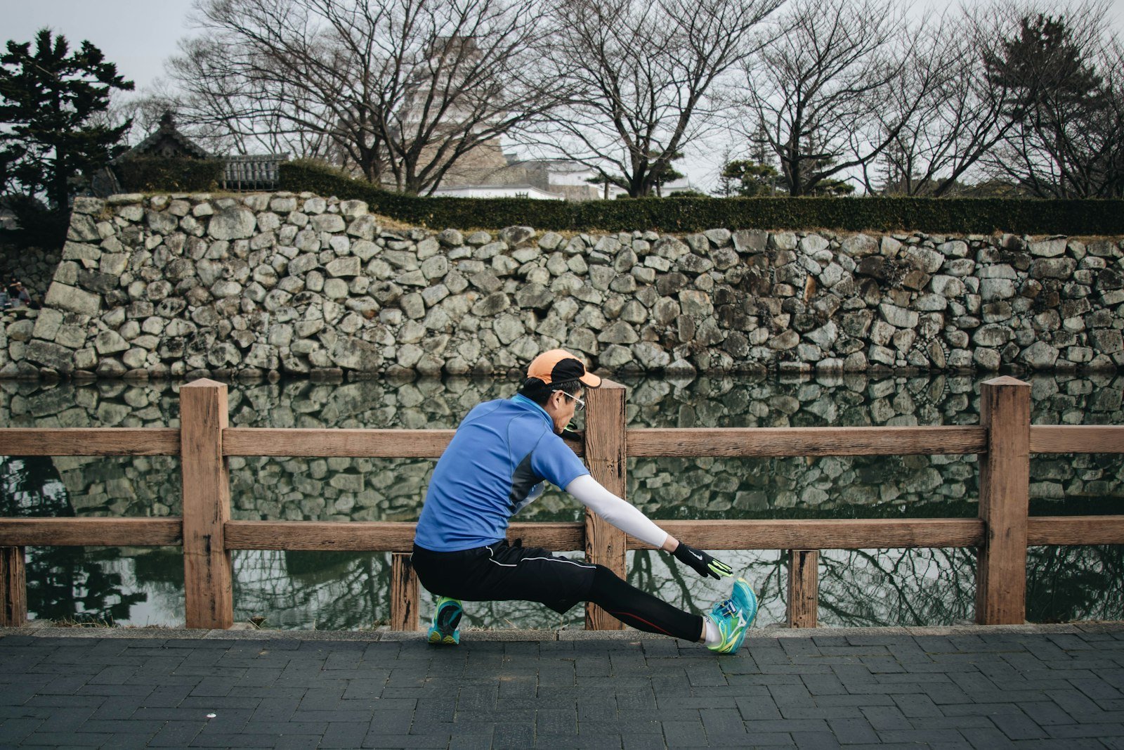Man in a blue top and black tights stretching against a wooden fence