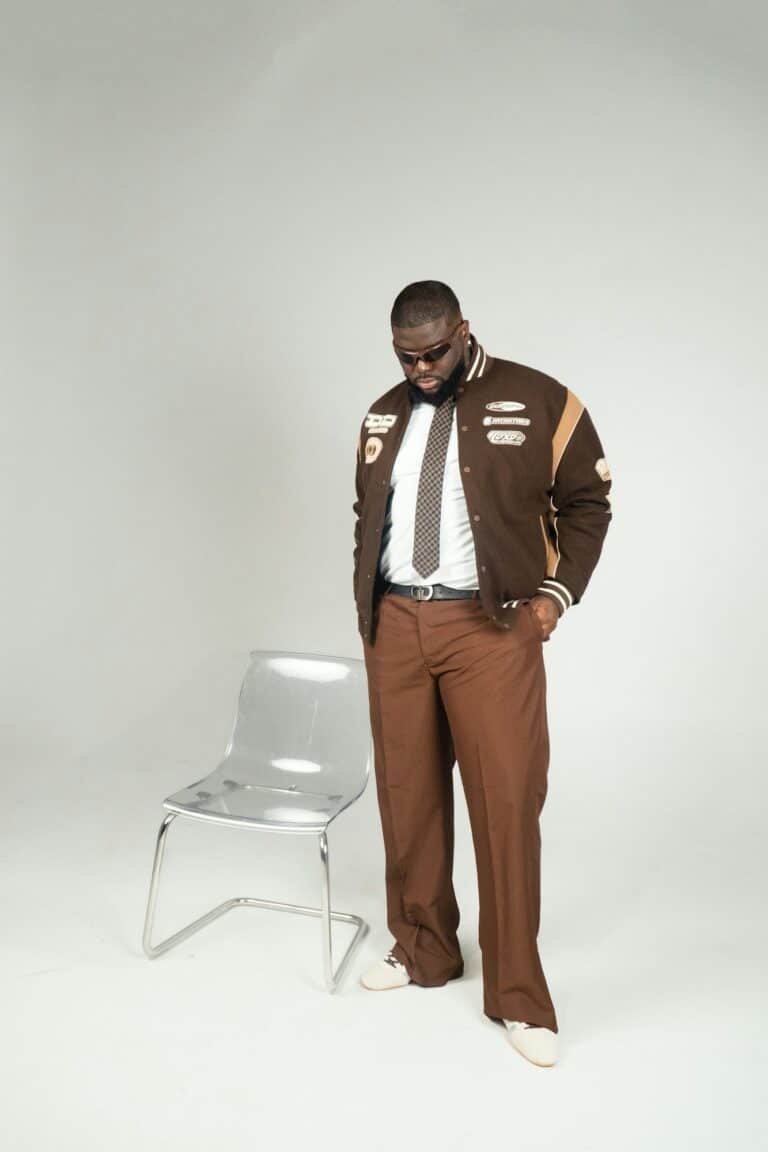 Man in a brown jacket and brown trousers standing by a chair