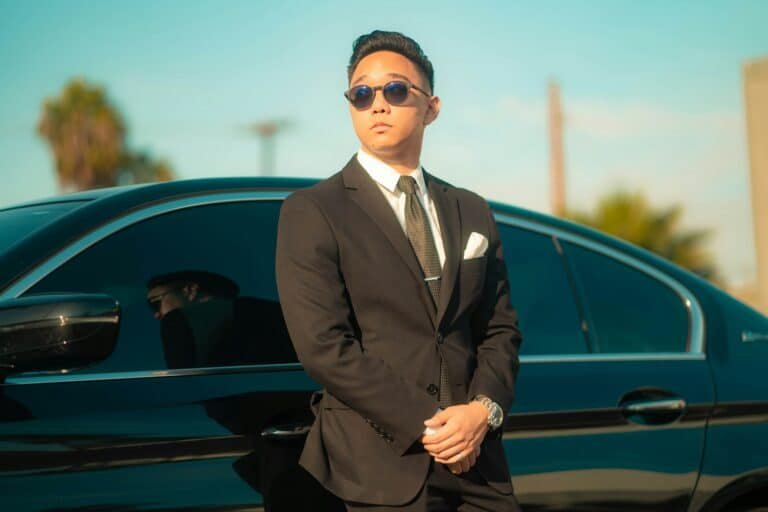 Man in a dark suit and sunglasses standing beside a car under palm trees