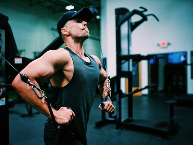 Man in a dark tank top and cap doing a cable exercise in a gym