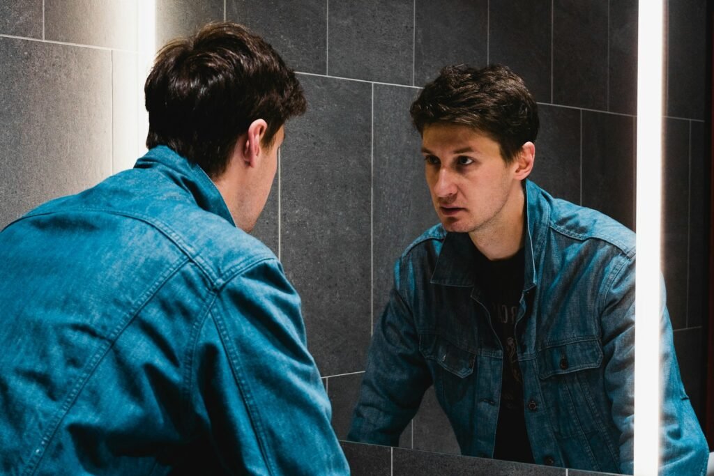 Man in a denim jacket studying his reflection in a bathroom mirror
