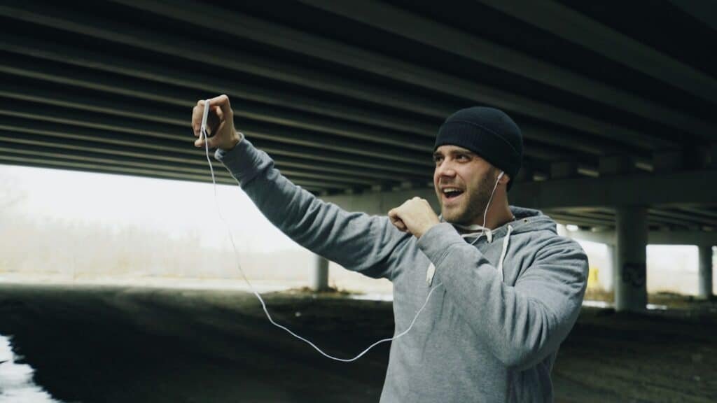 Man in a gray hoodie and beanie under a bridge, holding up his phone