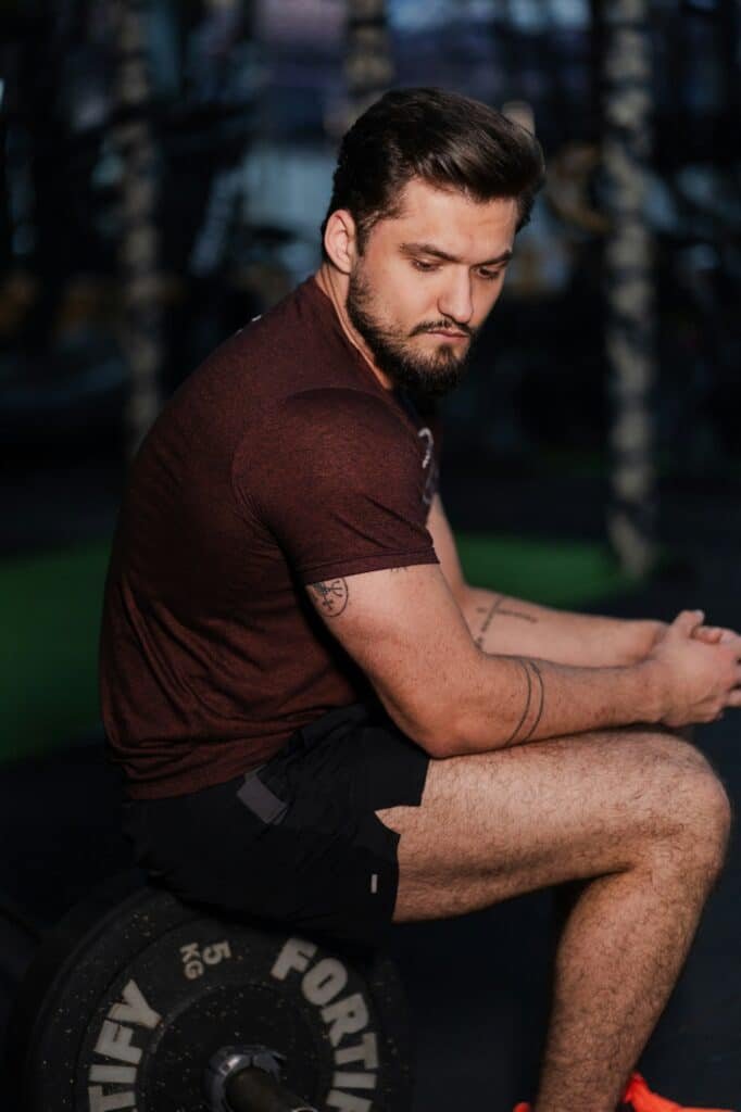 Man in a maroon tee and black shorts sitting on a weight plate in a gym