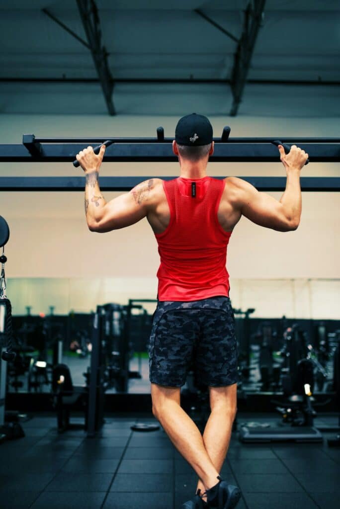 Man in a red tank top doing pull-ups in a gym, seen from behind