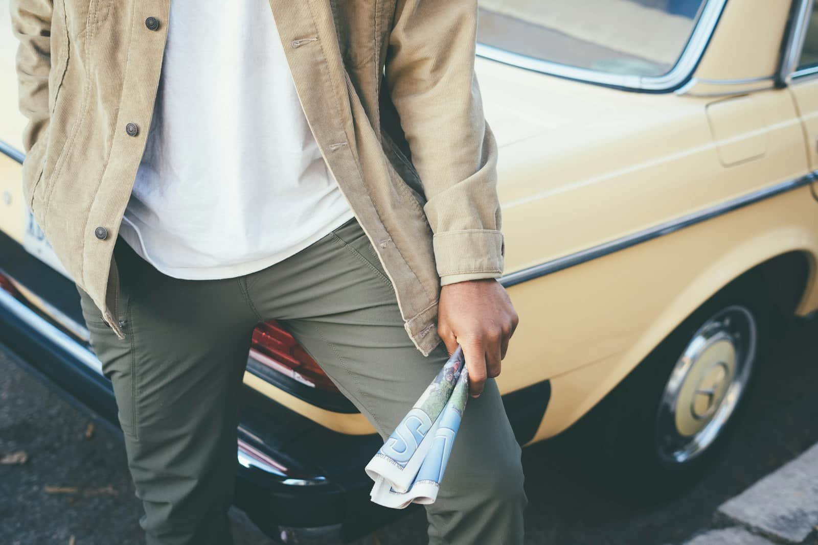 Man in a tan jacket, white tee and olive pants holding a newspaper beside a vintage car