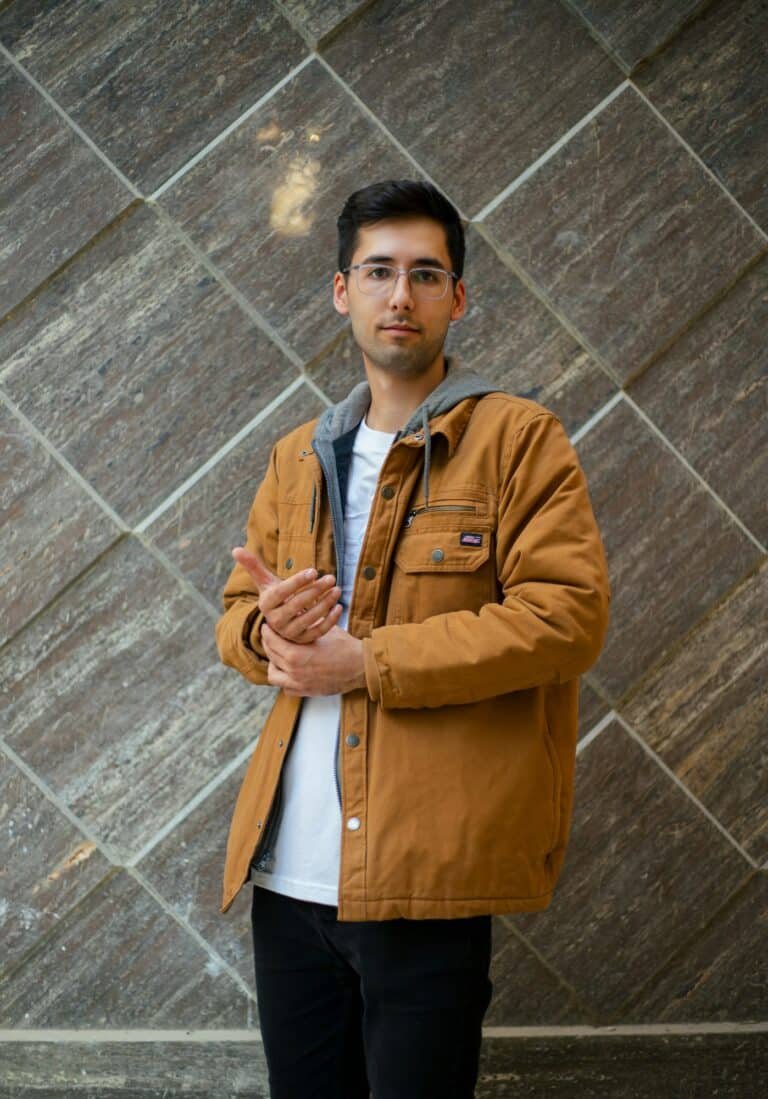Man in a tan work jacket over a white tee in front of a stone wall