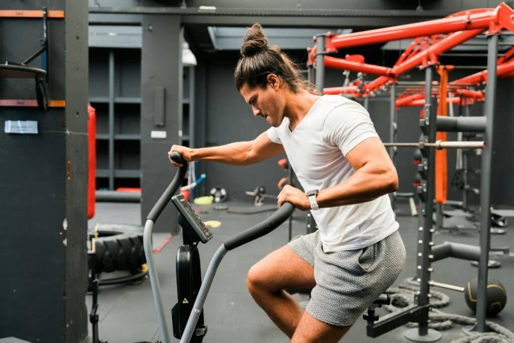 Man in a white tee and gray shorts working out on an air bike in a gym