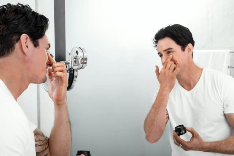 Man in a white tee holding a small jar and checking his skin in a bathroom mirror
