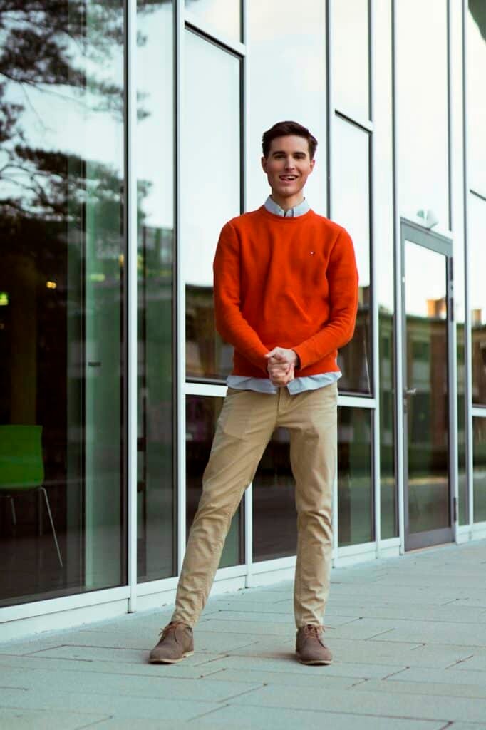 Man in an orange sweater and khaki trousers outside a glass building