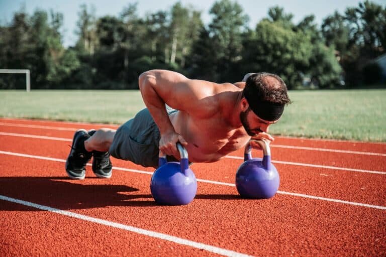 Man in gym shorts doing push-ups on two kettlebells on a running track
