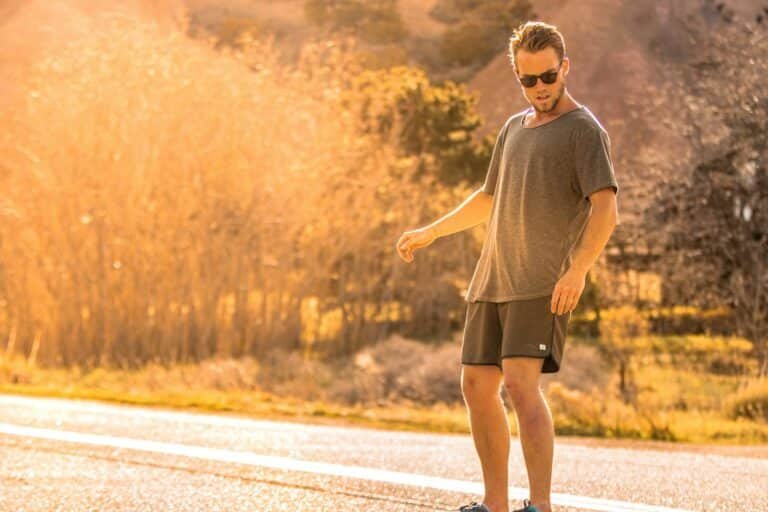 Man in sunglasses, a gray tee and dark shorts walking along a sunlit road