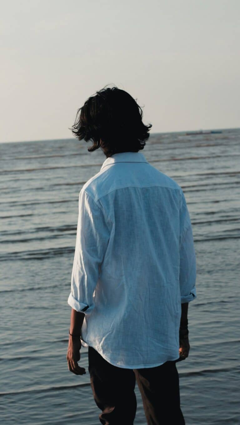 Man in a light blue linen shirt and dark trousers facing the sea
