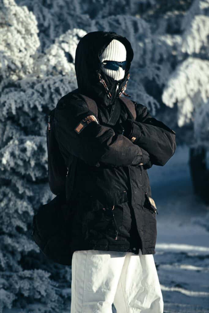 Person in a black winter jacket with their face covered, standing in snow
