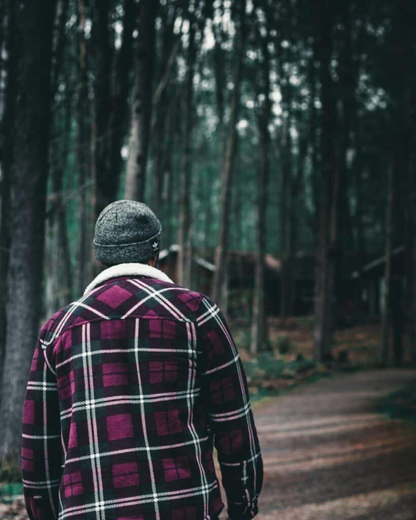 Man in a red, black and white plaid shirt and a grey knit cap walking down a forest road, seen from behind