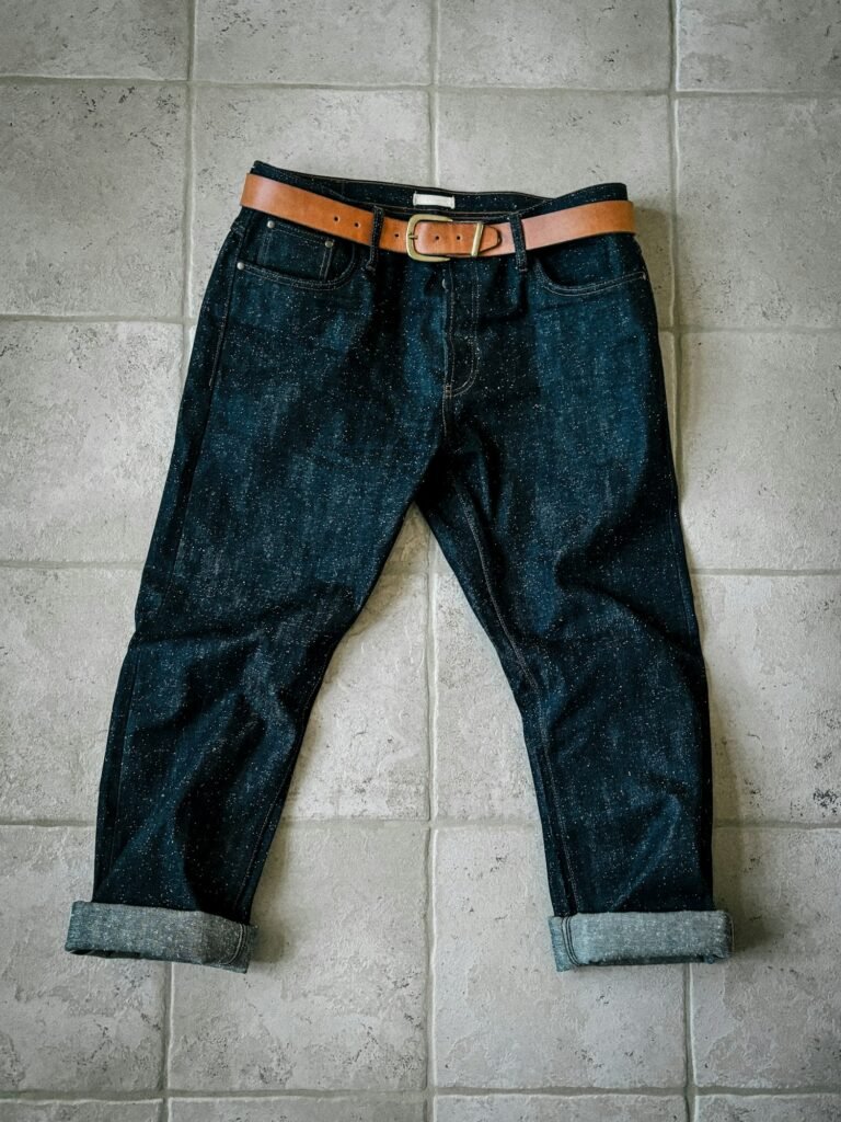 Dark raw denim jeans with a brown leather belt laid flat on a tile floor