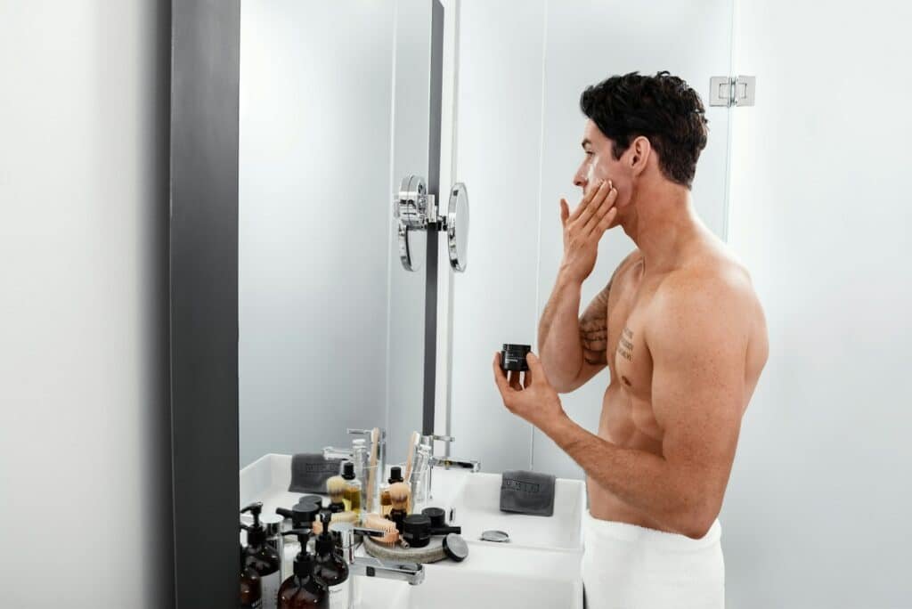 Shirtless man applying cream to his face at a bathroom mirror