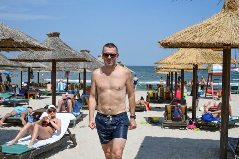 Shirtless man in navy swim trunks walking on a beach with straw umbrellas