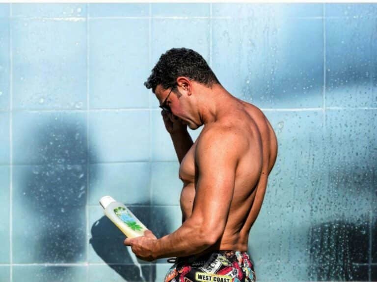 Shirtless man in swim trunks reading the label on a bottle by a tiled wall