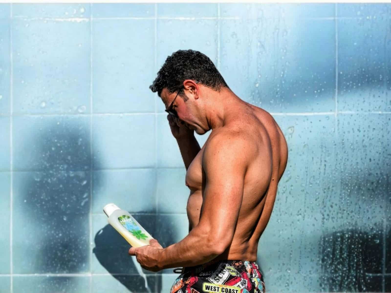 Shirtless man in swim trunks reading the label on a bottle by a tiled wall
