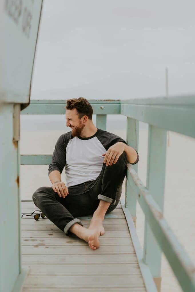 Smiling man in a raglan tee and dark pants sitting on a wooden deck