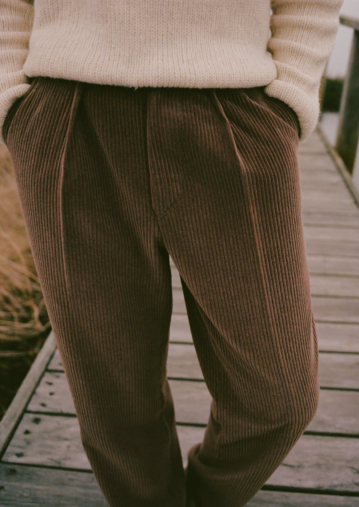 Pleated brown corduroy trousers with a cream ribbed sweater on a wooden dock