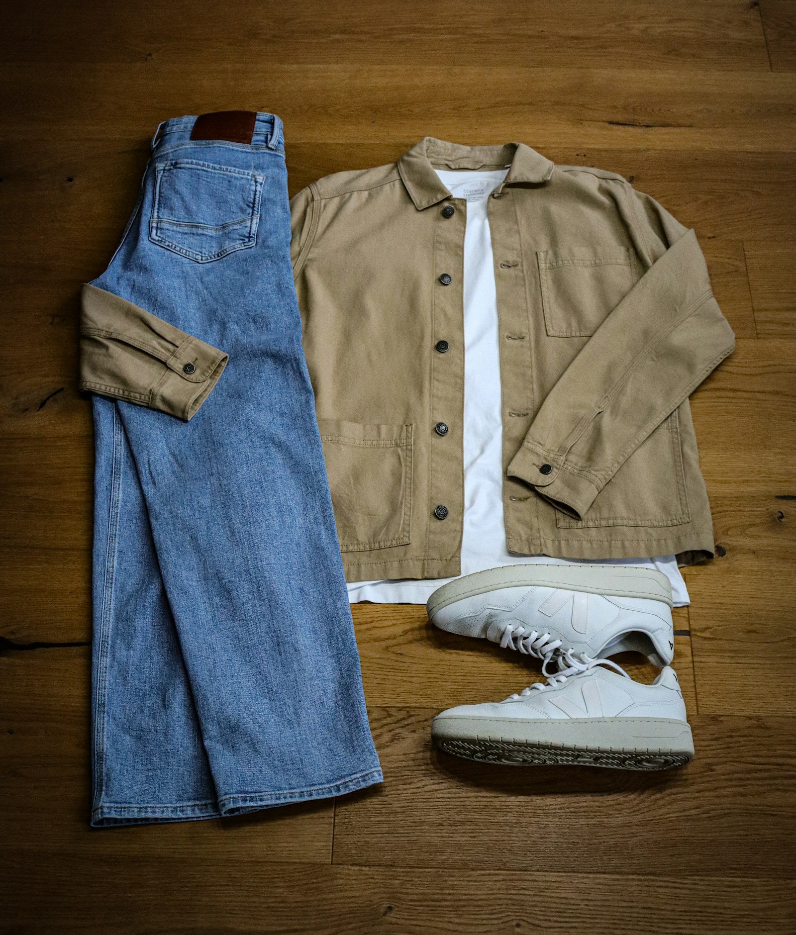Jeans, a tan chore jacket over a white tee and white sneakers laid flat on a wood floor
