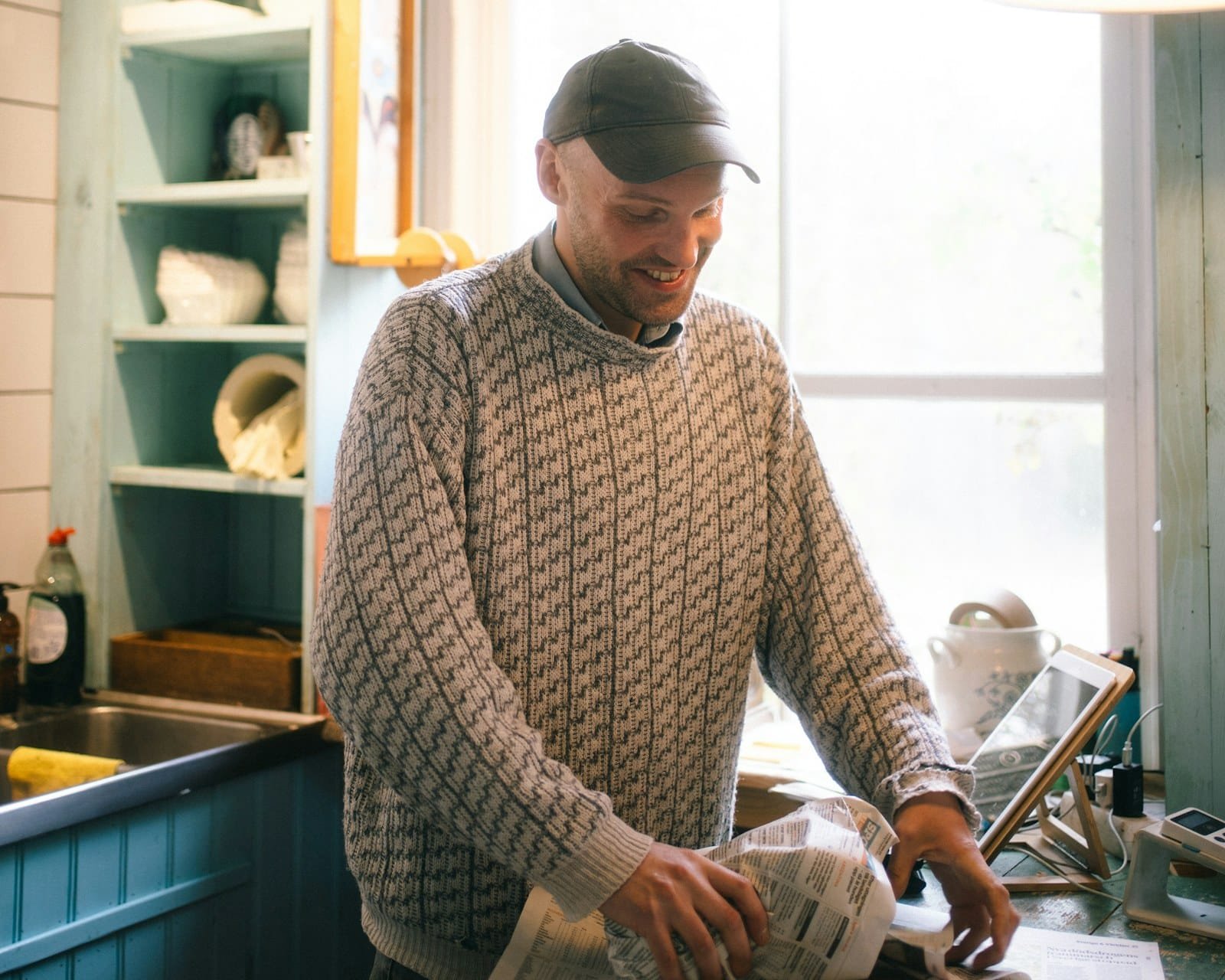 Man in a patterned knit sweater wrapping something in newspaper at a kitchen counter