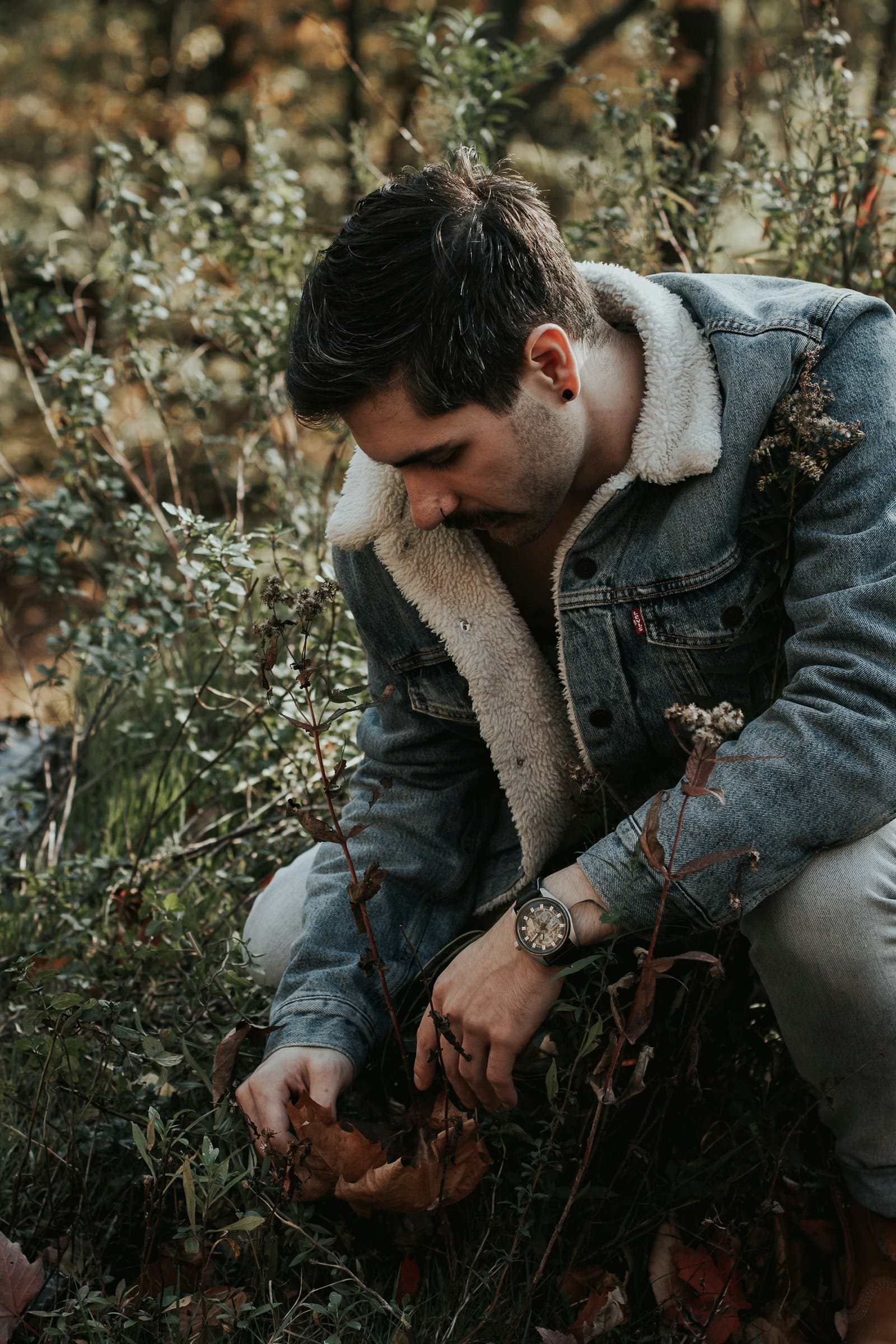 Man in a sherpa-lined denim jacket crouching among autumn plants